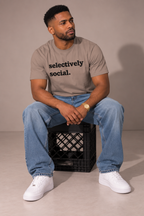 A man sitting on a black milk crate wearing a gray graphic t-shirt with the text 'selectively social.' in black, blue jeans, and white sneakers, set against a plain studio backdrop.
