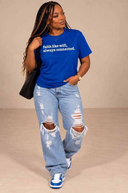 A woman modeling a bright blue crewneck t-shirt with the white printed text 'faith like wifi, always connected.' She is wearing distressed light-wash blue jeans with ripped knees, blue and white sneakers, and carrying a black shoulder bag against a solid beige studio background.