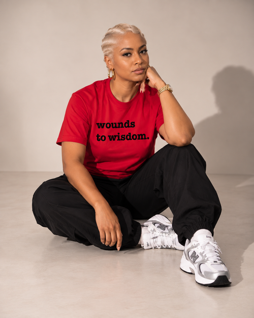 A model sits cross-legged on a light gray studio floor wearing a bright red graphic t-shirt with the black text 'wounds to wisdom.' and black sweatpants with white and gray New Balance sneakers.